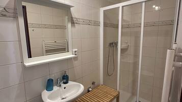 A bathroom with a toilet, sink, mirror, shower, and a wooden shelf.