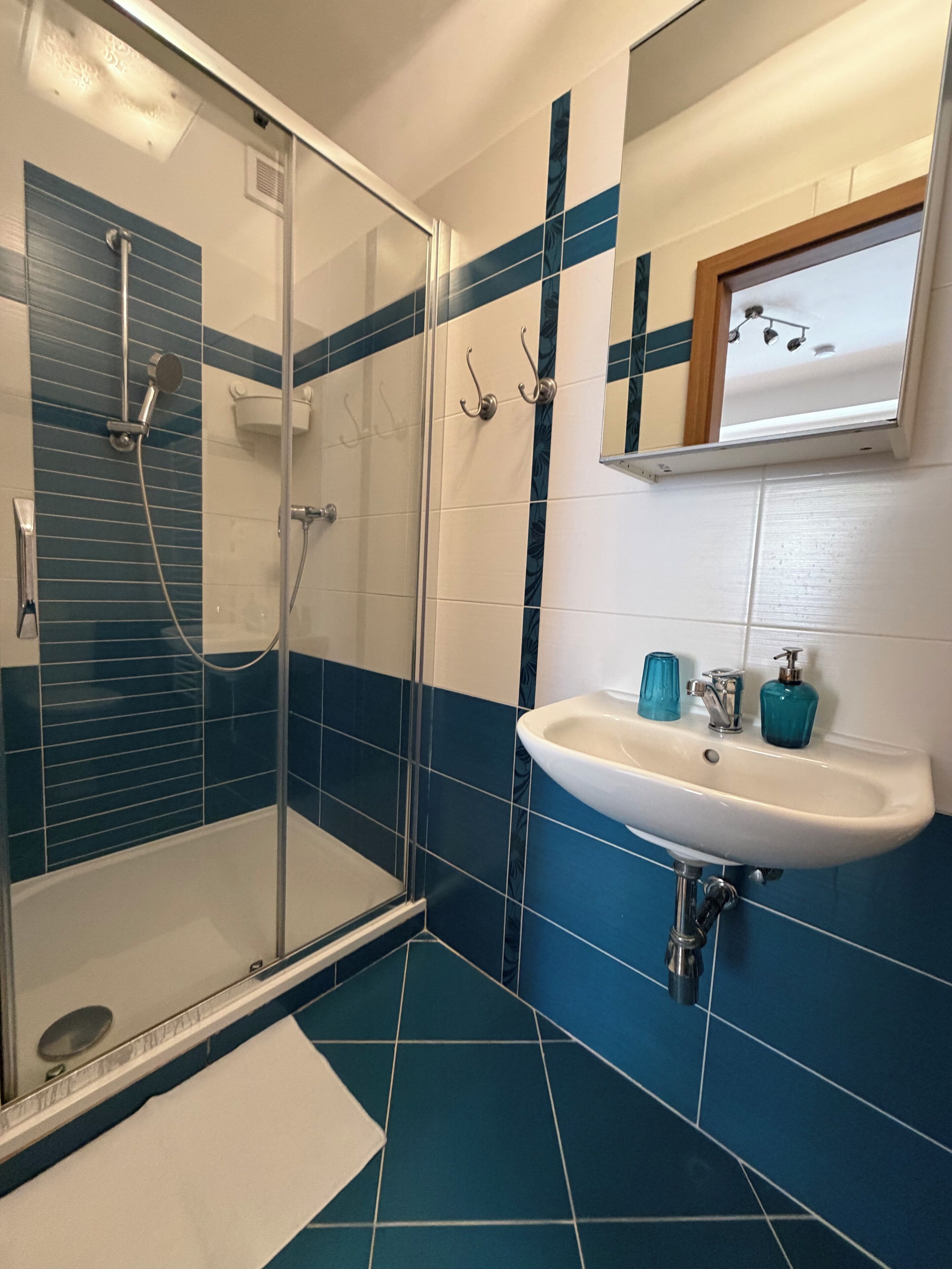 A bathroom with a glass shower enclosure, a white sink with blue soap dispensers, and a mirror with a wooden frame.