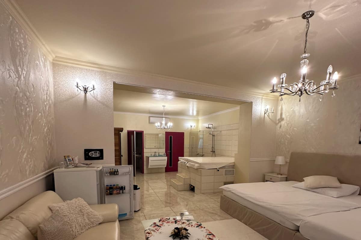 Une chambre d’hôtel avec un lit, un canapé, une petite table recouverte d’une nappe à motifs floraux, un lustre, et une salle de bain visible au fond.