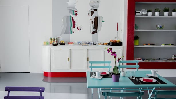 A modern kitchen with white cabinets, a red accent wall, and a dining area with a teal table and chairs. - B&B il Castelluccio (Arezzo)