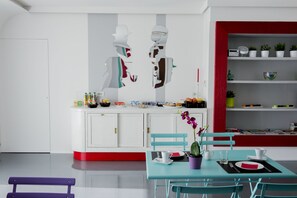 A modern kitchen with white cabinets, a red accent wall, and a dining area with a teal table and chairs. - B&B il Castelluccio (Arezzo)