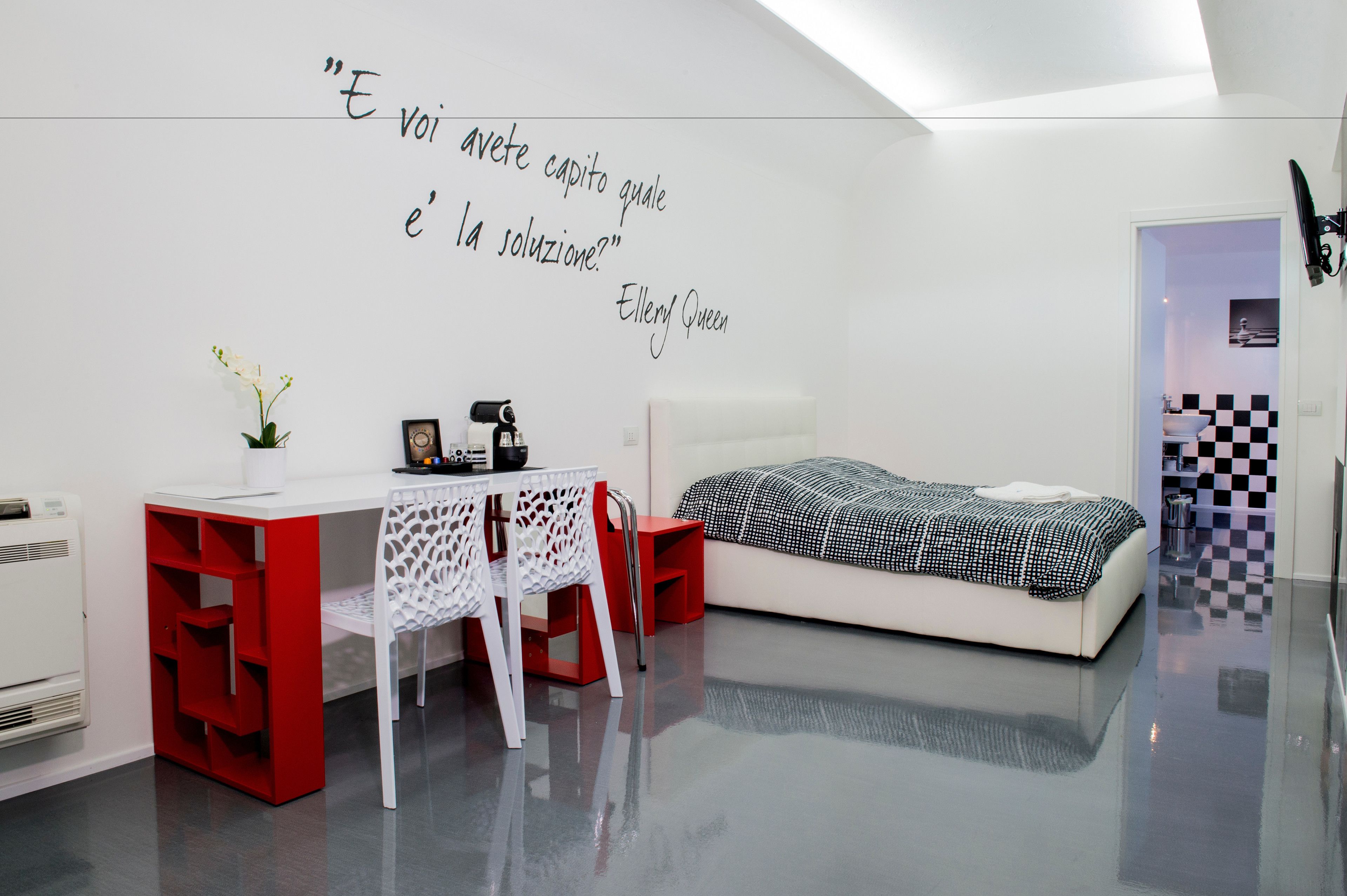 A modern bedroom with a white bed, a desk with a red chair, and a wall quote by Ellery Queen.