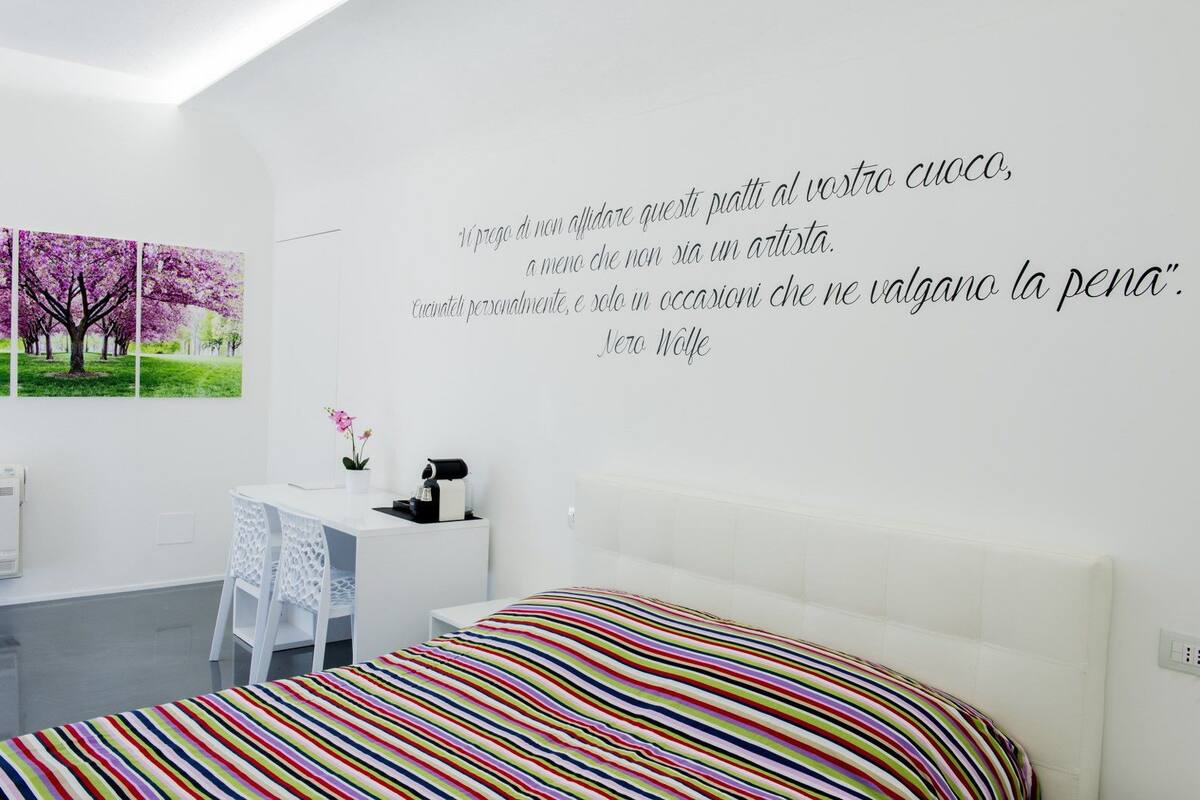 A modern bedroom with a white bed, colorful striped bedding, a desk with a chair, and a wall with a quote in Italian.