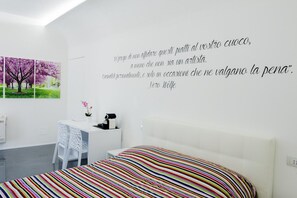 A modern bedroom with a white bed, colorful striped bedding, a desk with a chair, and a wall with a quote in Italian. - B&B il Castelluccio (Arezzo)