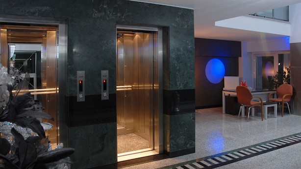 Un ascensor en un vestíbulo moderno con piso de mármol y una zona de estar con sillas naranjas.
