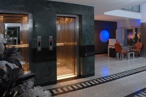 An elevator in a modern lobby with a marble floor and a seating area with orange chairs.