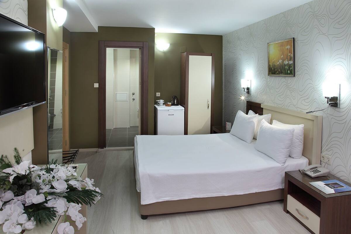 Habitación de hotel con una cama grande, una mesita de noche con lámpara, un televisor montado en la pared y una puerta que da a un baño.
