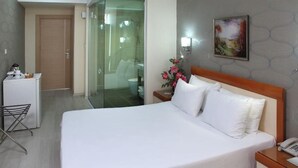 Habitación de hotel con cama, mesita de noche, un escritorio con teléfono, una silla, un refrigerador pequeño, cabina de ducha de vidrio y un cuadro en la pared.
