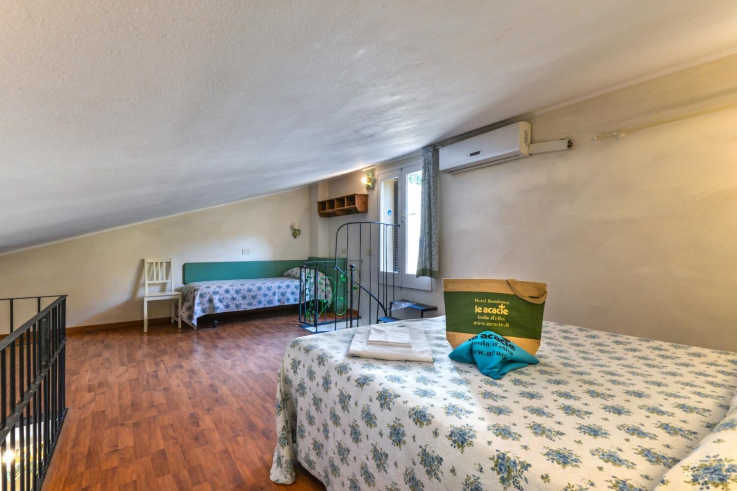 A small, attic-style room with a wooden floor, two single beds with floral bedding, a chair, and a table.
