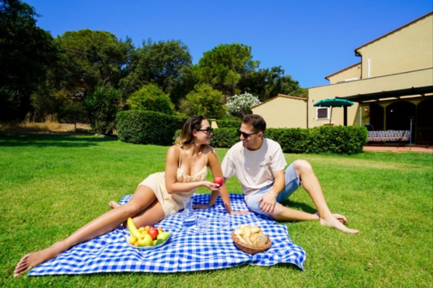 Two people sitting on a checkered blanket outdoors with a table of fruits and bread.