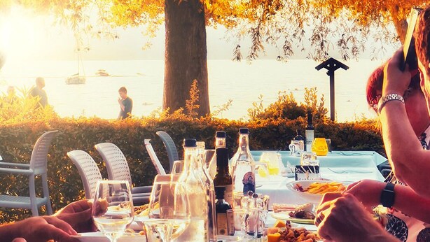 Mesa de comedor al aire libre con diferentes platos y bebidas, con vistas a un cuerpo de agua.