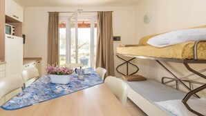 Una habitación compacta con literas, una mesa de comedor con sillas, una pequeña cocina y una ventana con cortinas.