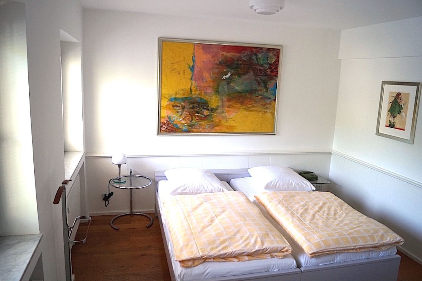 A hotel room with two beds, a painting on the wall, and a small table with a lamp.