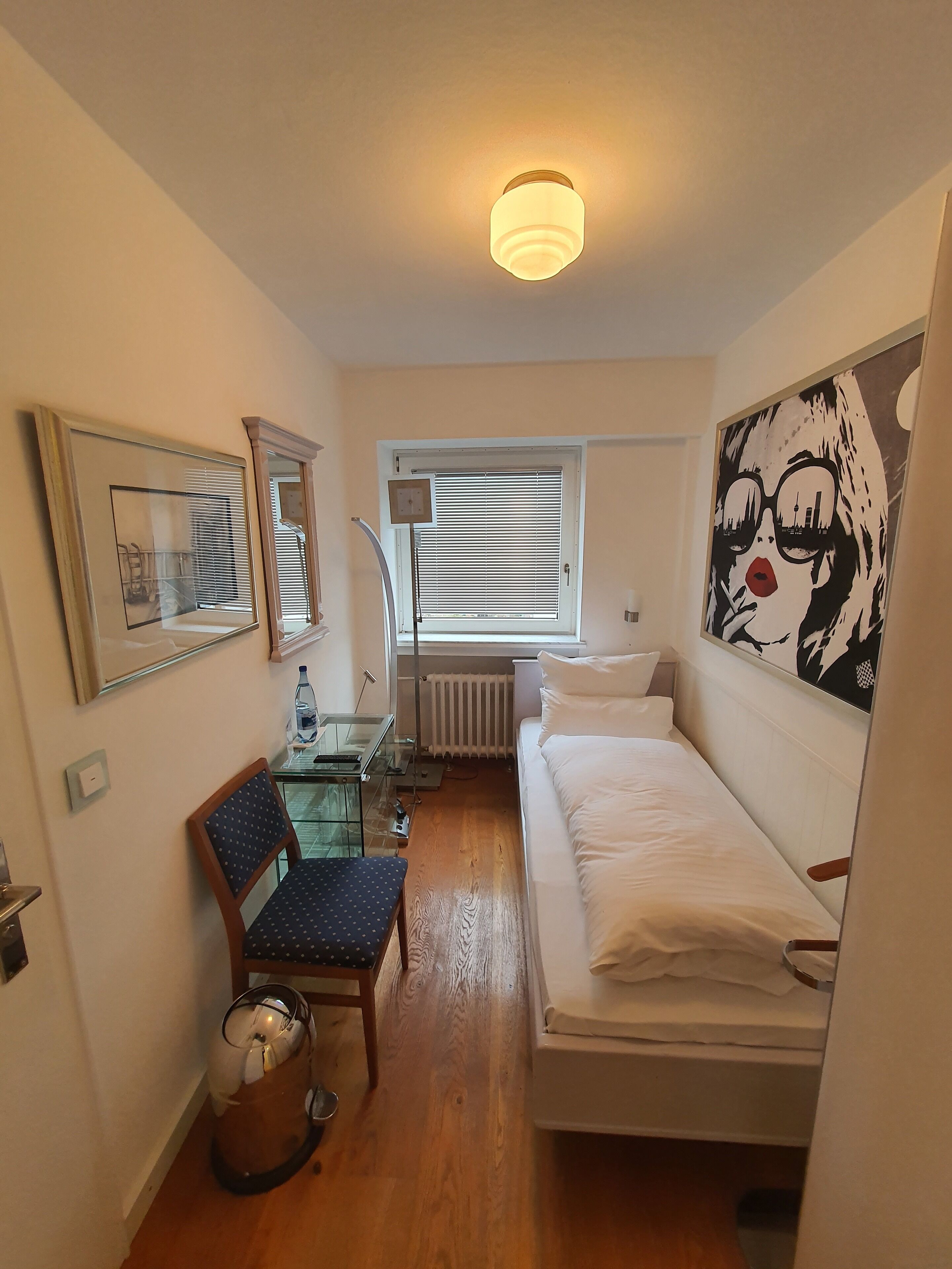 A small, well-lit room with a bed, a desk with a chair, a radiator, and a window with blinds.