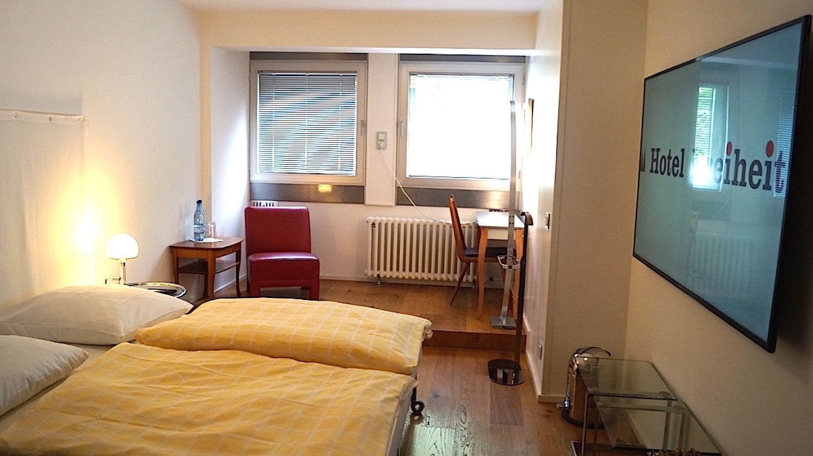 A hotel room with two beds, a desk, a chair, a radiator, and a TV on the wall.