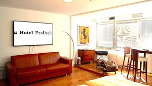 A living room with a red sofa, a small table with a lamp, a chair, and a bed. There is a large window with blinds and a picture frame on the wall. - Hotel Freiheit (Cologne)