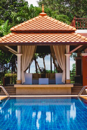 Un área de piscina con una estructura de techo con tejas, un espacio de descanso con cojines y una piscina azul cristalina.
