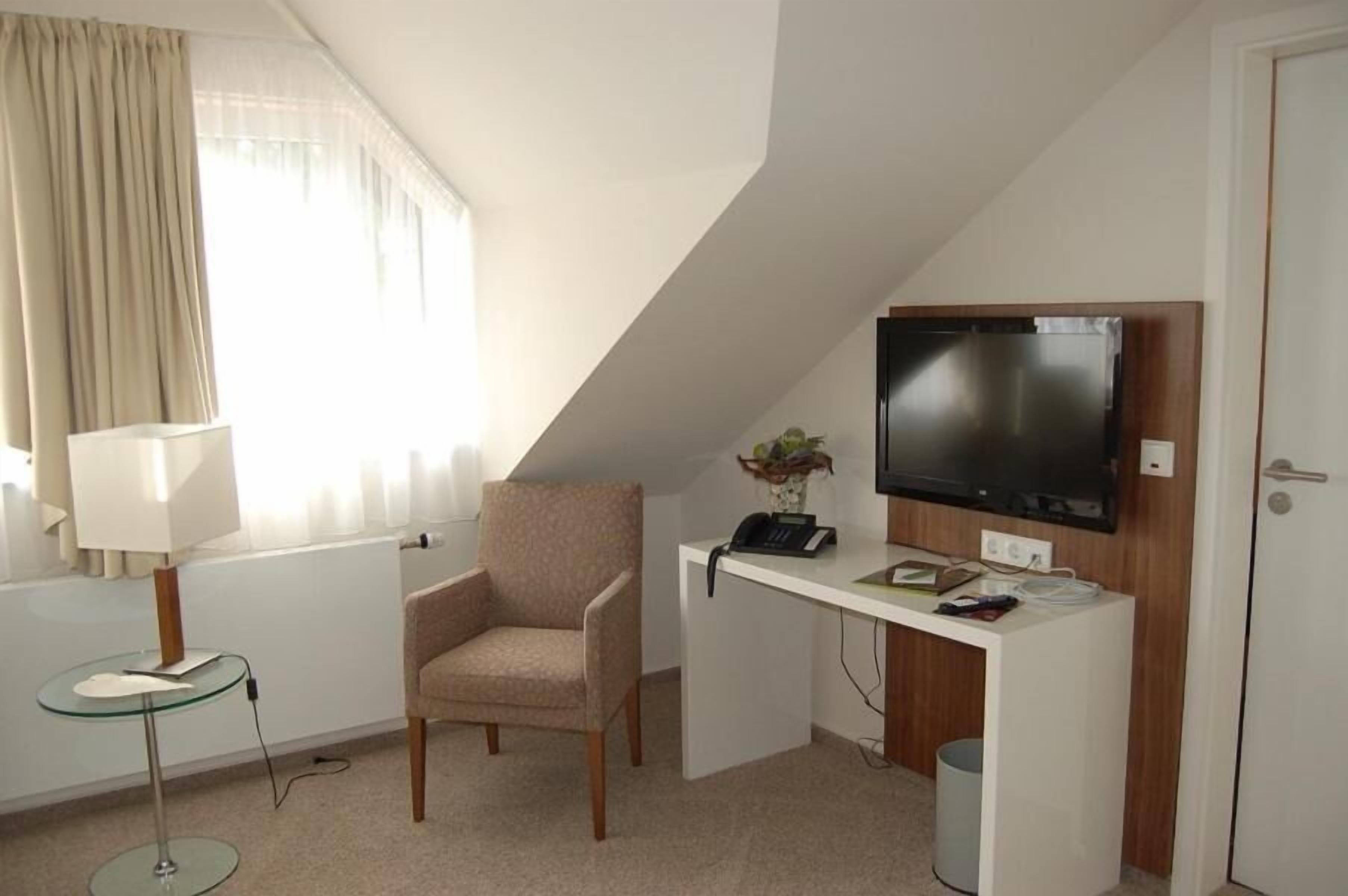 A hotel room with a desk, chair, television, lamp, and a window with curtains.