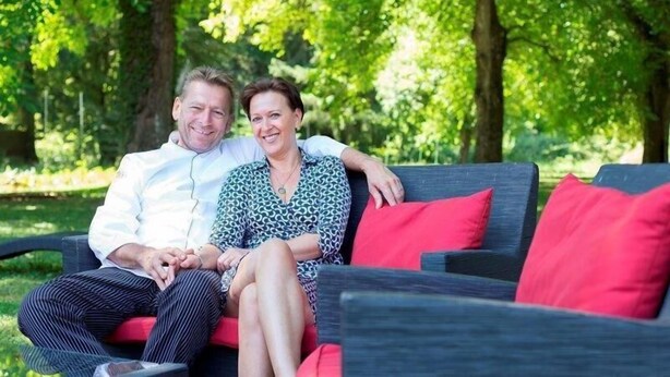 A couple sitting on a dark-colored sofa with red cushions in a lush green outdoor setting. - Restaurant Niedmühle Land & Genuss Hotel (Rehlingen-Siersburg)
