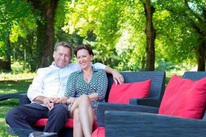A couple sitting on a dark-colored sofa with red cushions in a lush green outdoor setting.