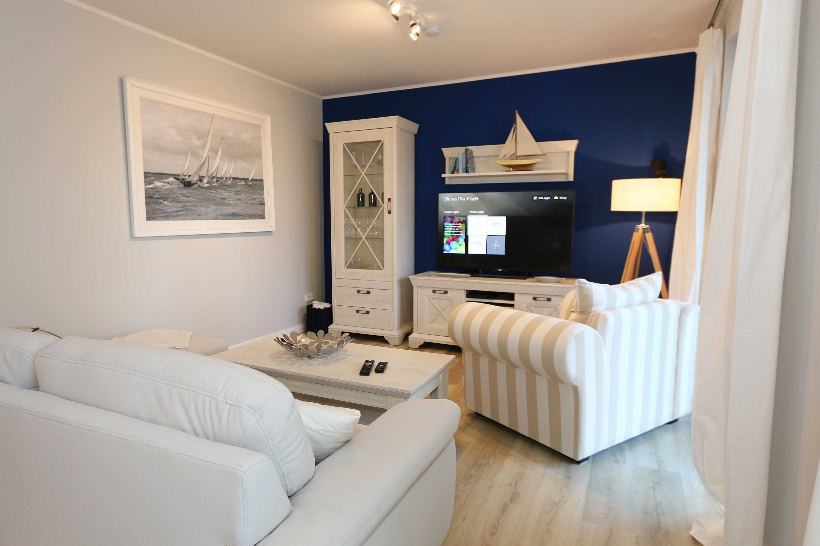 Una sala de estar con un sofá blanco, un sillón a rayas, un televisor, un mueble bajo y un cuadro en la pared.
