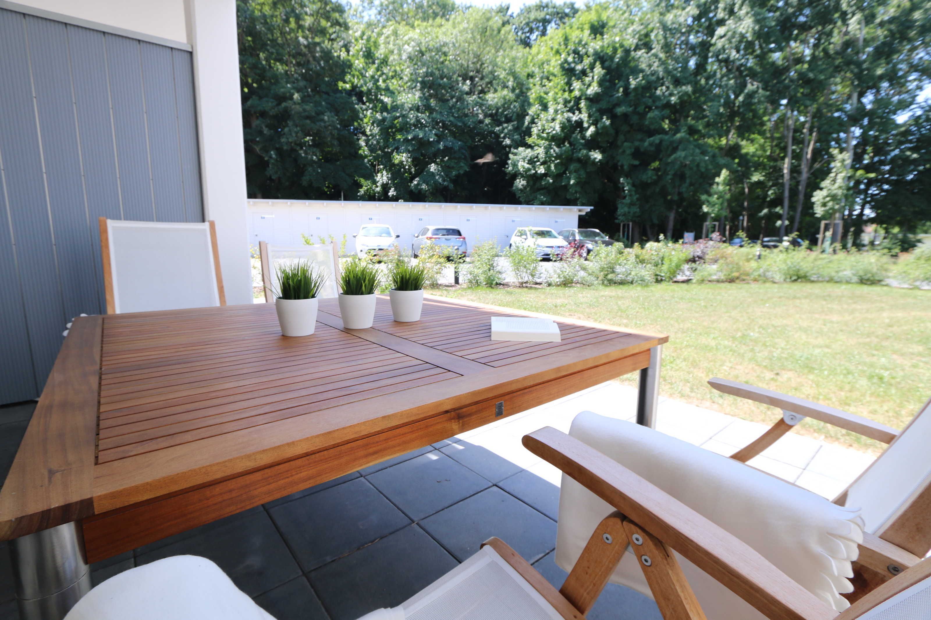 Une table en bois, entourée de chaises et agrémentée de plantes en pot, est placée sur une terrasse d’où l’on peut admirer un parking et des arbres.