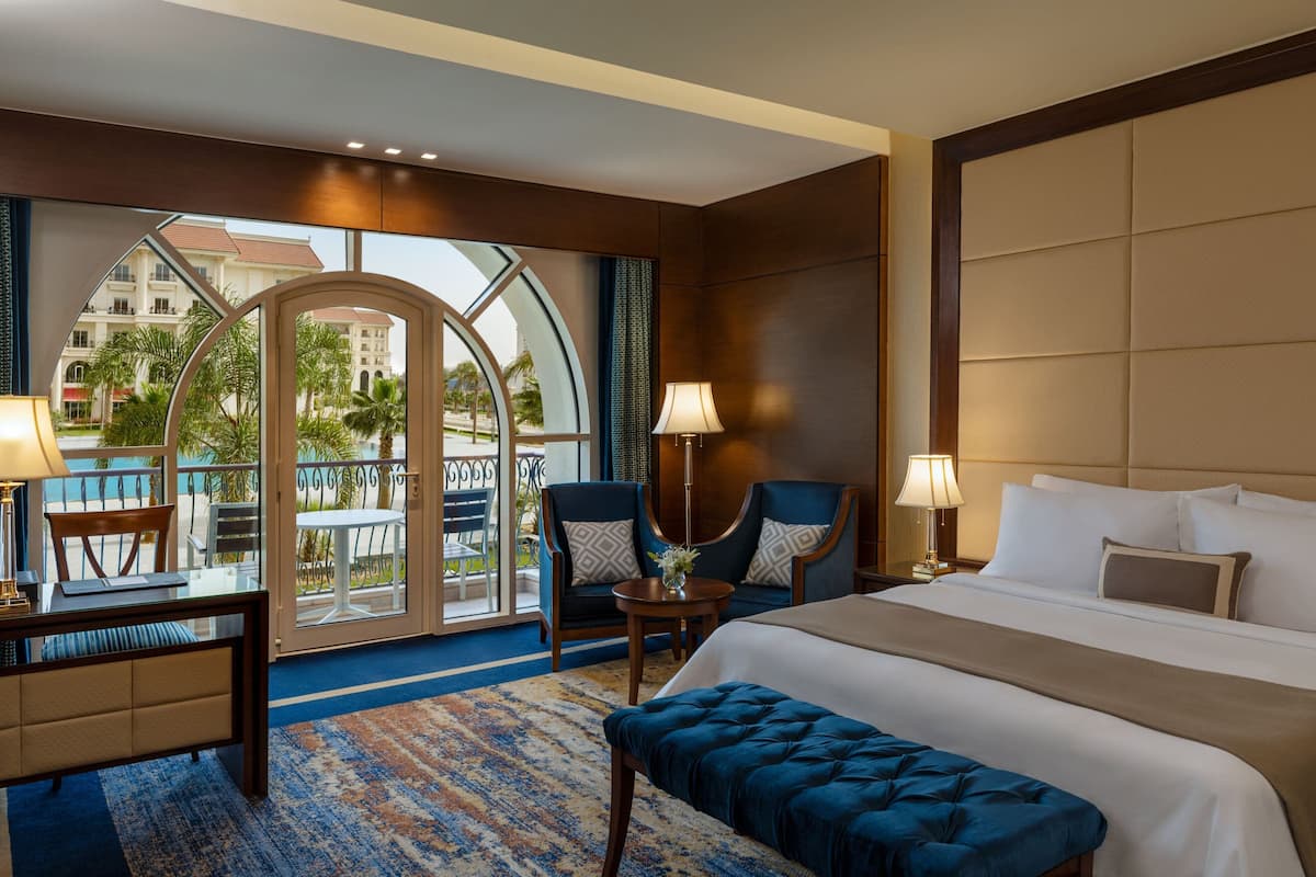 A hotel room with a large bed, two armchairs, a desk, and a view of a building and palm trees through a glass door.
