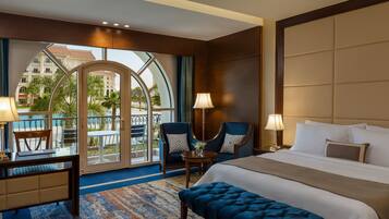 A hotel room with a large bed, two armchairs, a desk, and a view of a building and palm trees through a glass door.
