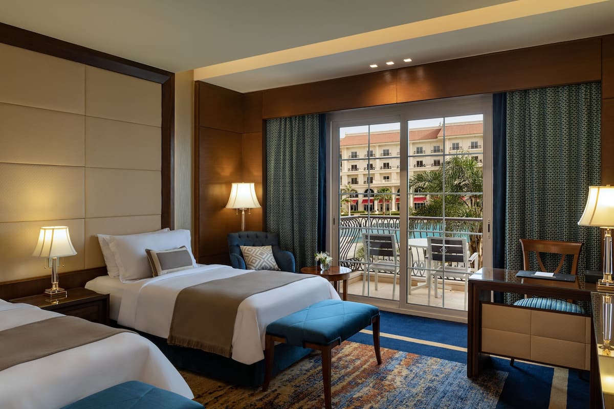 A hotel room with two beds, a desk, and a balcony with a view of buildings.