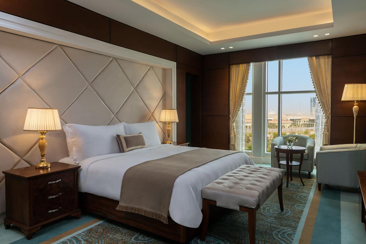 A hotel room with a large bed, two bedside lamps, a small table, a chair, and a view of the city through large windows.