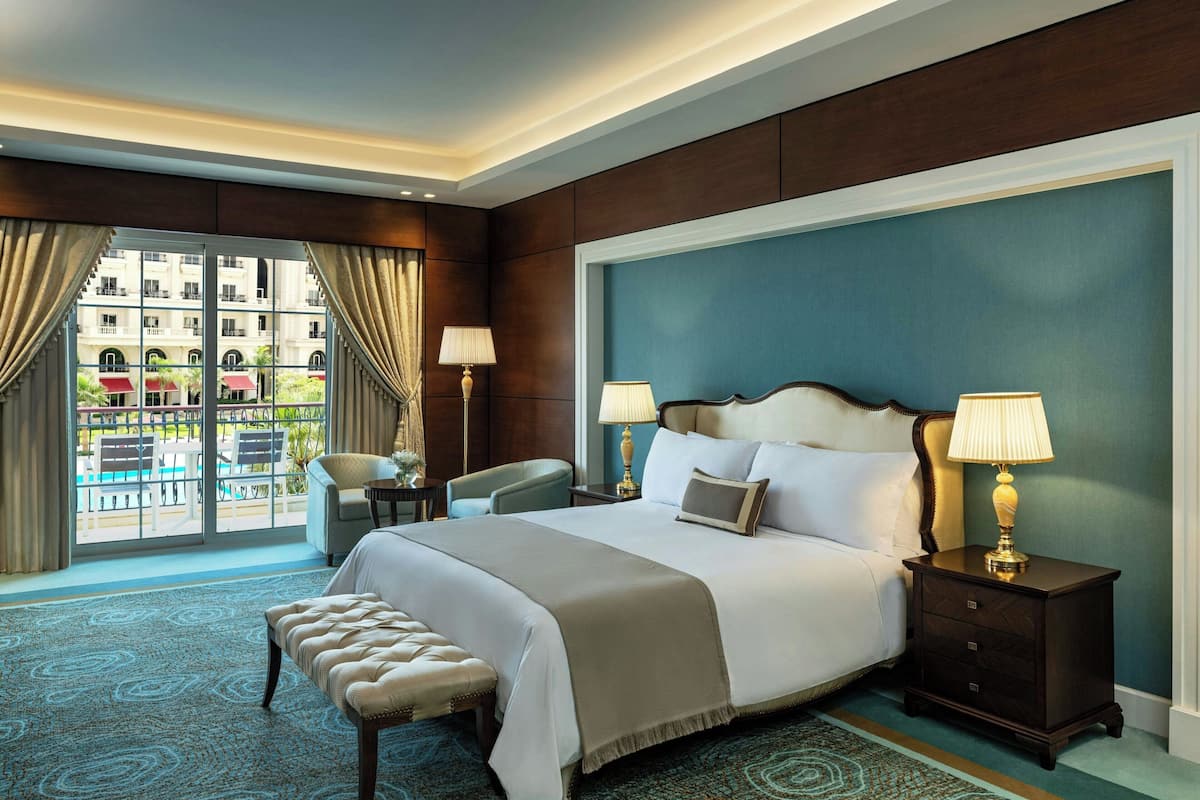 A hotel room with a large bed, two bedside tables with lamps, a bench, and a view of a pool and outdoor seating area through large windows.