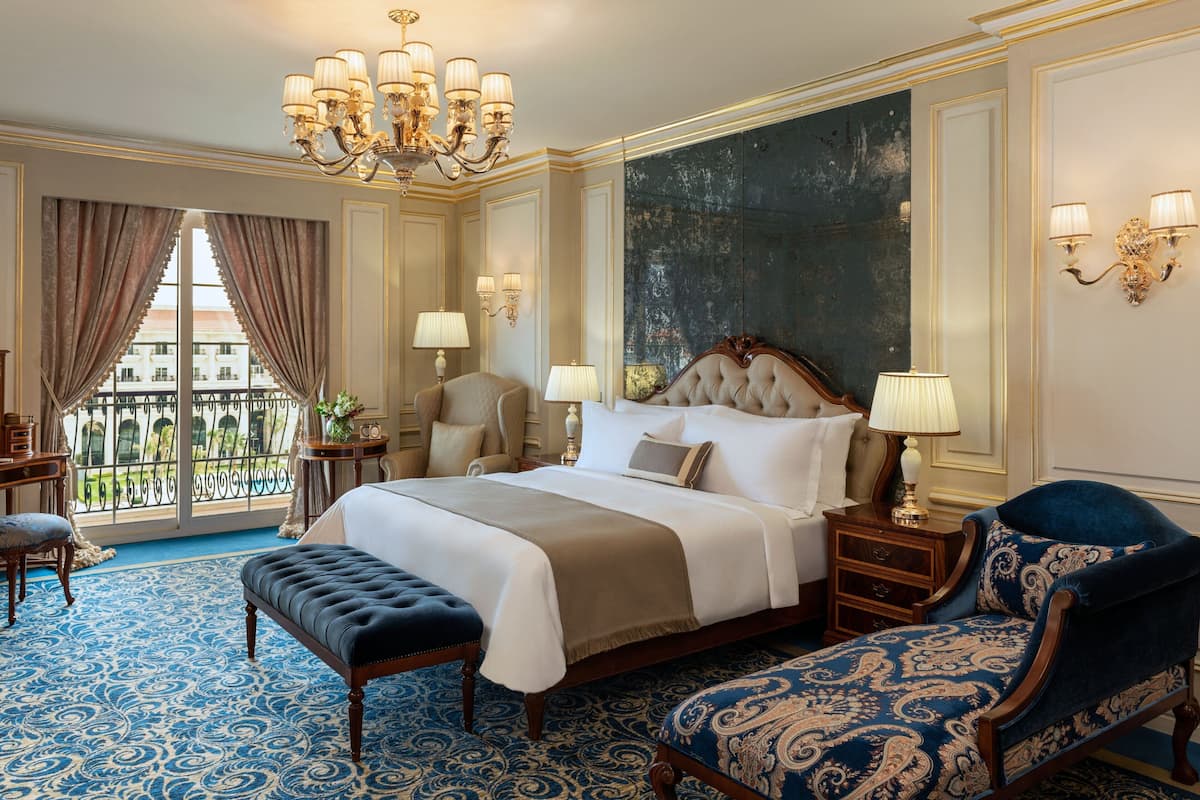 A luxurious bedroom with a large bed, a chaise lounge, a wooden nightstand, and a chandelier.