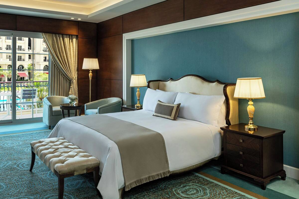 A hotel room with a large bed, two bedside tables with lamps, a bench, a view of the city through a large window, and a balcony with outdoor seating.