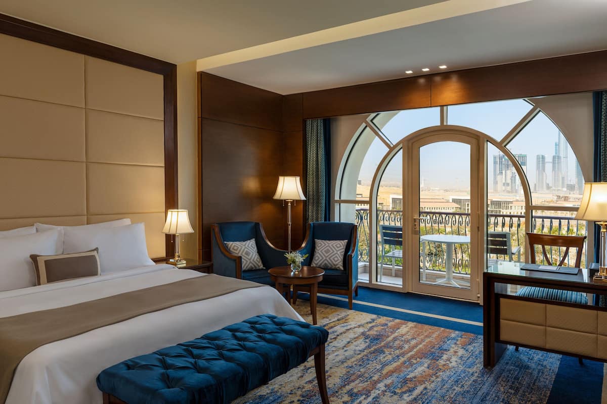 A hotel room with a large bed, two armchairs, a desk, and a view of a cityscape through a large window.