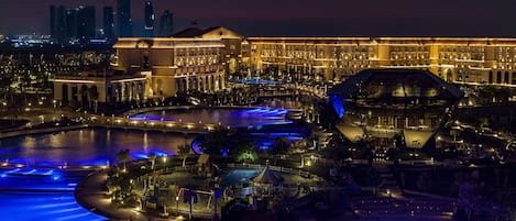 A large hotel with a central courtyard, multiple swimming pools, and a city skyline in the background.
