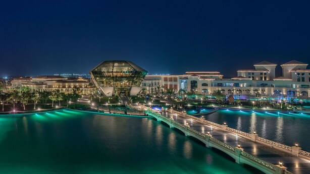 A waterfront hotel with a distinctive glass structure, a pool area, and a bridge. - The St. Regis New Capital, Cairo (New Capital)