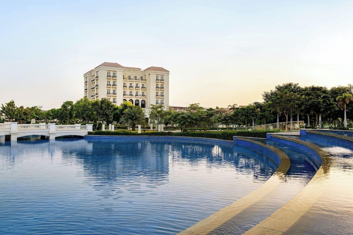 A large swimming pool with a bridge, surrounded by greenery and a building in the background.