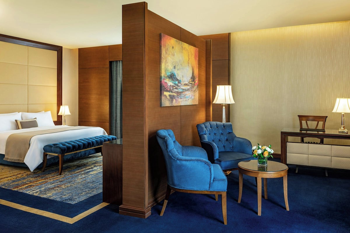 A hotel room with a bed, a blue armchair, a small table with a vase, and a painting on the wall.