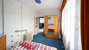 A bedroom with a bed, a wardrobe, a small bedside table, a mirror, and a door leading to another room.