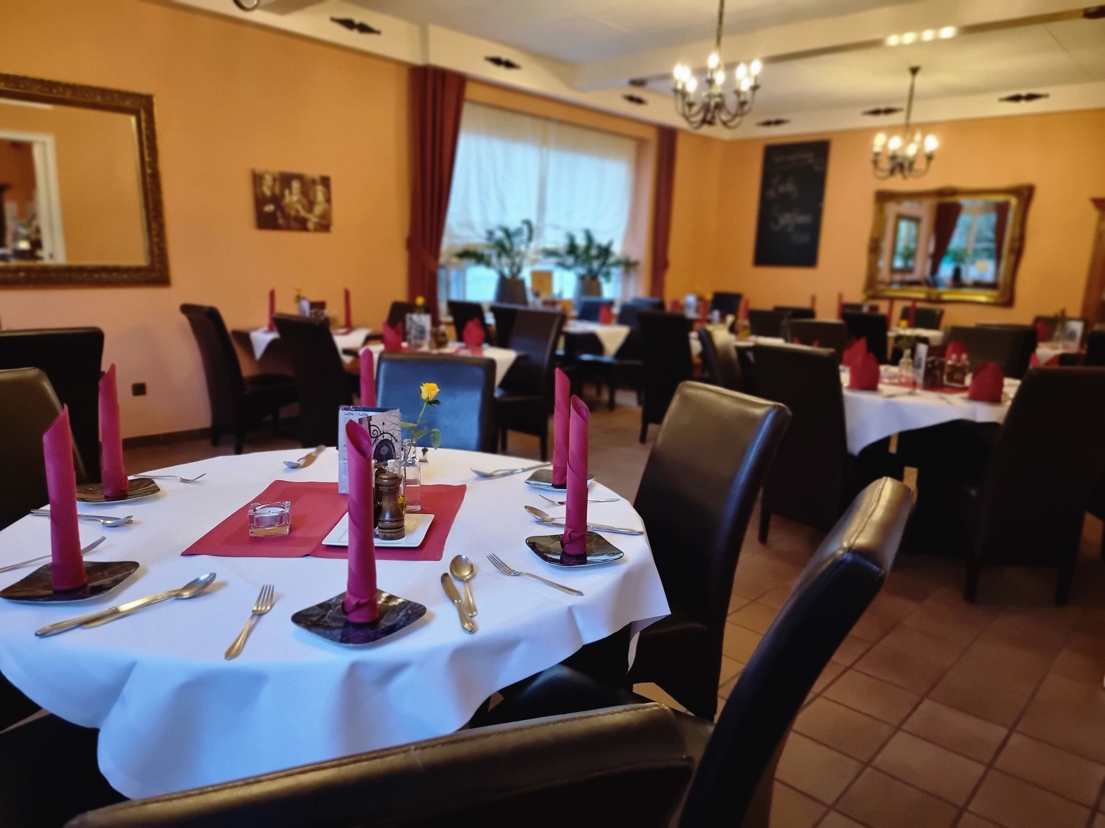 Ein Essbereich mit runden Tischen für zwei Personen, jeweils mit weißer Tischdecke, roten Servietten und goldfarbenem Besteck.