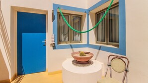 Une pièce avec une porte bleue, un piédestal blanc sur lequel repose un bol en terre cuite contenant un cactus, et une chaise en osier.