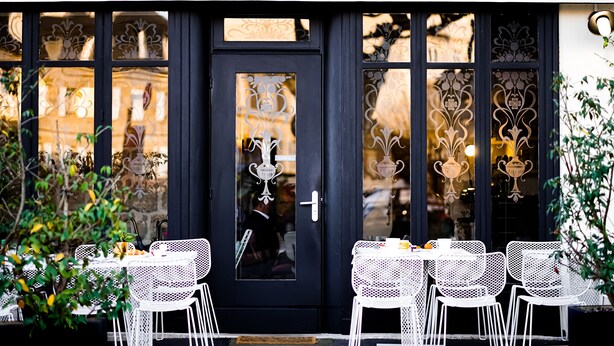 A hotel entrance with a dark door, white metal chairs, and tables set up outside. - Hôtel Le Collonges (Brive-la-Gaillarde)