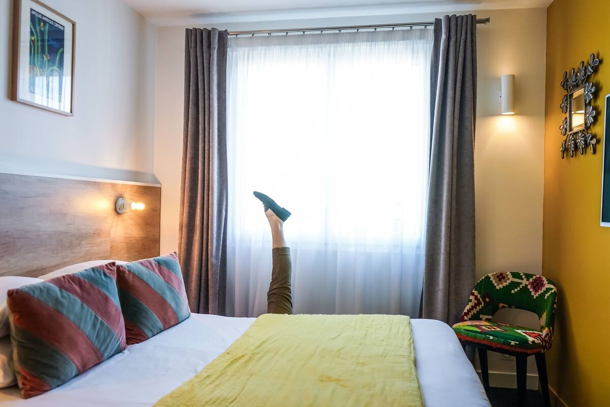 A hotel room with a bed, a chair, a window with curtains, and a wall-mounted light fixture.