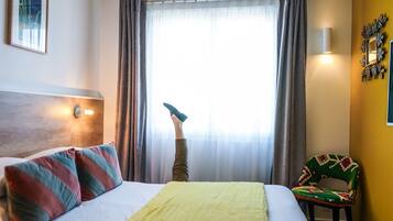 Una habitación de hotel con cama, una silla, una ventana con cortinas y una lámpara fijada a la pared.