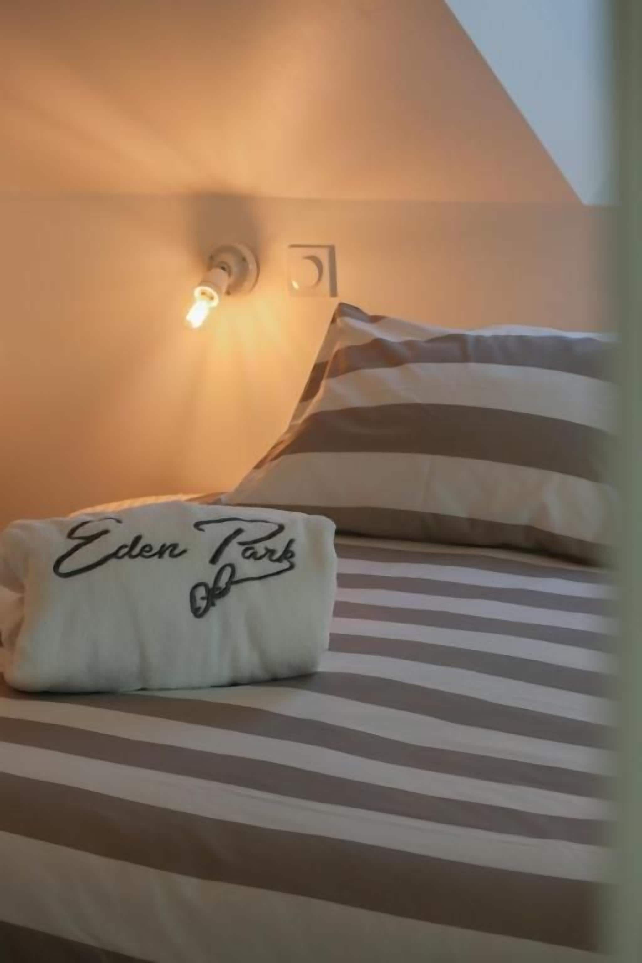 Una cama con ropa de cama a rayas, un almohadón con el texto 'Eden Park' y una lámpara de pared.