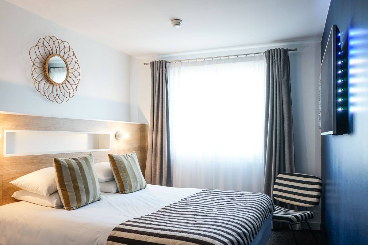 A hotel room with a large bed, a bedside table, a mirror, a chair, and a window with curtains.
