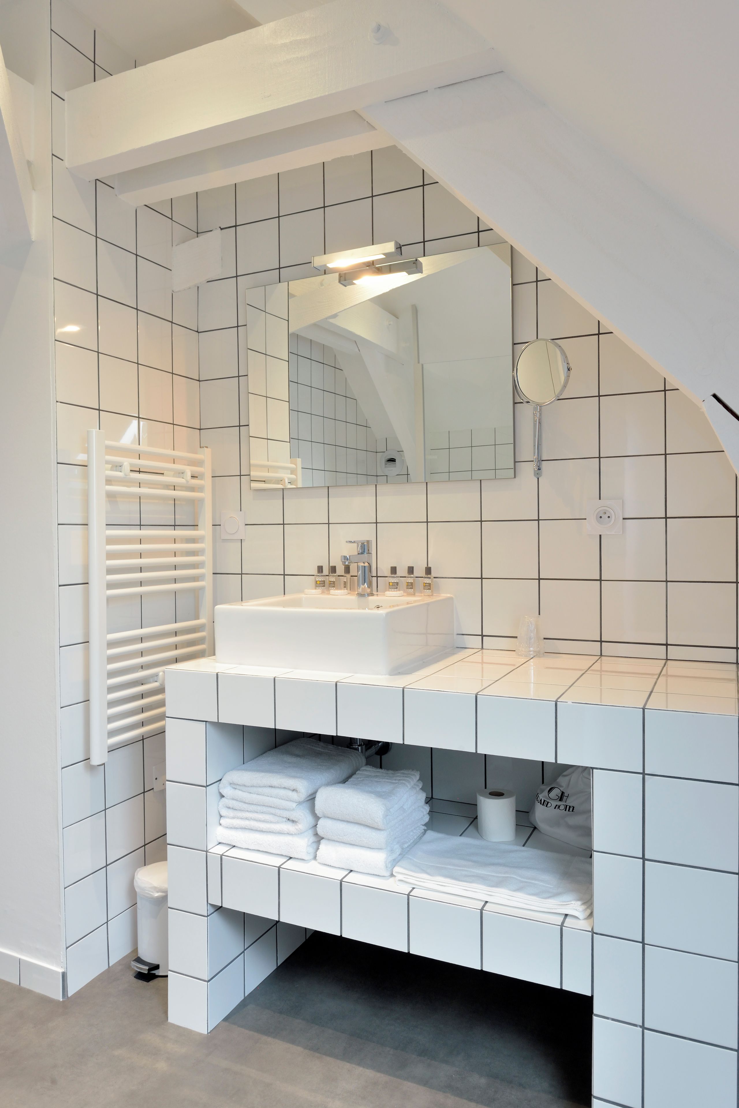 Un baño moderno con pared de azulejos blancos, un espejo grande, un lavamanos blanco y un perchero para toallas.