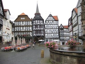 Une place de ville européenne traditionnelle, avec des bâtiments à colombages, des terrasses et une fontaine.