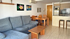 A modern living room with a grey sofa, wooden dining table, and chairs. There is a kitchen area with a microwave, stove, and cabinets.
