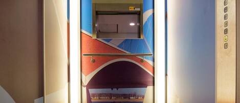 Un pasillo estrecho con un mural azul y rojo en las paredes, un ascensor y una alfombra con un símbolo blanco.
